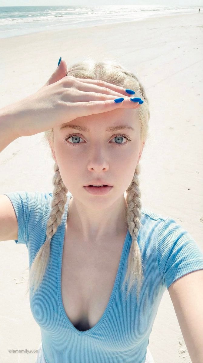 Ein High-Angle-Selfie-Foto einer jungen Frau mit Porzellanpuppen-Merkmalen an einem hellen Strand, die eine Hand zum Schutz der Augen vor das Gesicht hält, während sie zur Kamera aufblickt. Sie trägt ein hellblaues Oberteil und blauen Nagellack